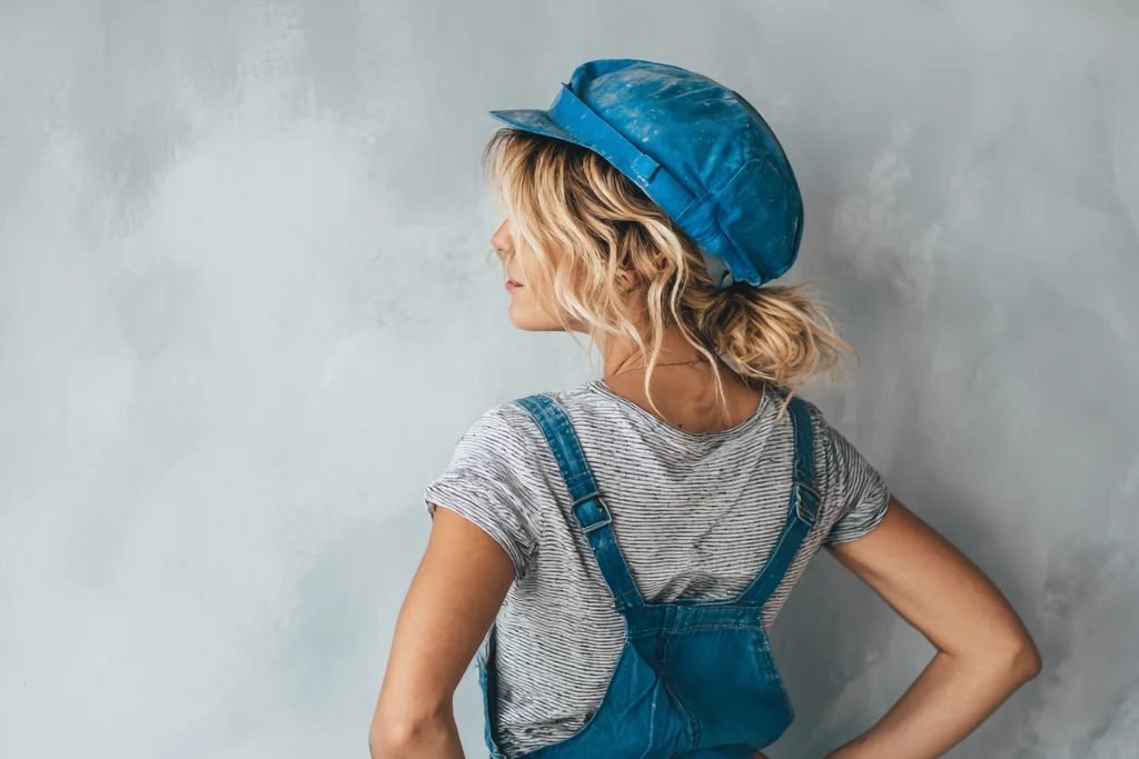 Malerin in blauer Latzhose und Schirmmütze vor einer frisch gespachtelten Wand