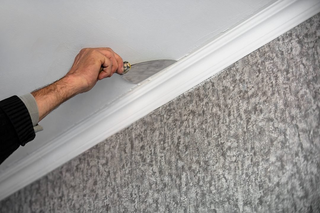Spachtel glättet die Wandfläche entlang einer weißen Zierleiste unter der Decke